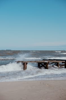 Storm sea waves empty wooden pier in dramatic blue sky background. Seascape with running splashing sea waves. Sandy beach vacation getaway in Gdansk Poland.
