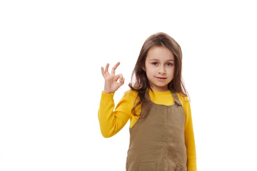 Caucasian confident smart schoolgirl showing OK sign, expressing hapiness and positive emotions over white background