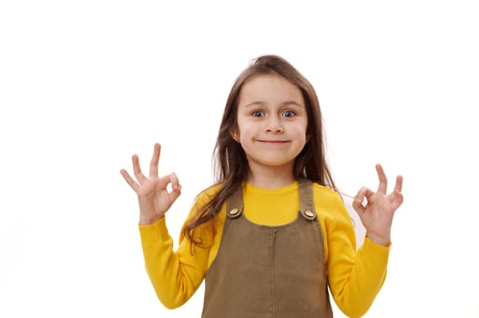Mischievous smiling little child girl showing OK sign, expressing hapiness and positive emotions over white background
