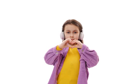 Happy little child girl in headphones, showing heart shape made from her fingers, expressing love and sympathy at camera
