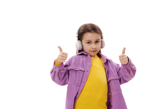 Elementary age child, lovely little school girl in wireless headphones, gestures with hands, shows thumbs up at camera