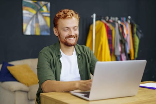 Caucasian man with red hair, sitting at desk in cozy home interior. He work on laptop, emphasizing concept of freelance work and ability to work from home. Man using his laptop to access his work remotely.