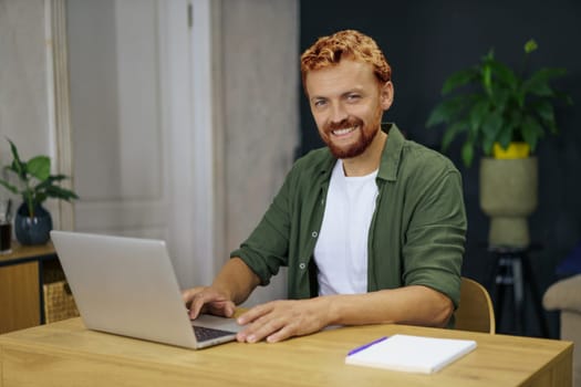 Caucasian man with foxy hair sitting at desk in a cozy and comfortable home interior. He is using laptop to work from home, emphasizing the concept of remote work and the benefits of flexibility and independence.
