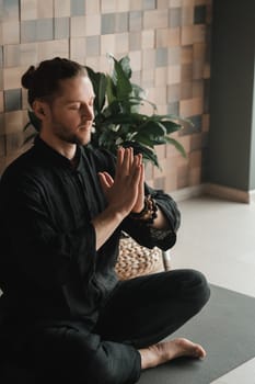 Portrait of a young man in a black kimano sitting in a lotus position on a gym mat in the interior
