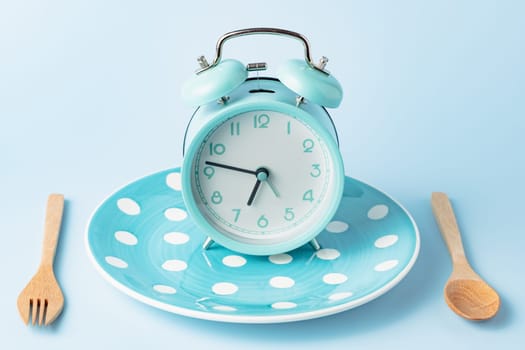 An alarm clock on an empty plate and cutlery set against blue background 