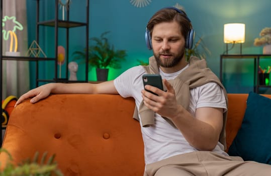 Happy caucasian man in wireless headphones relaxing sitting on sofa at home listening favorite music