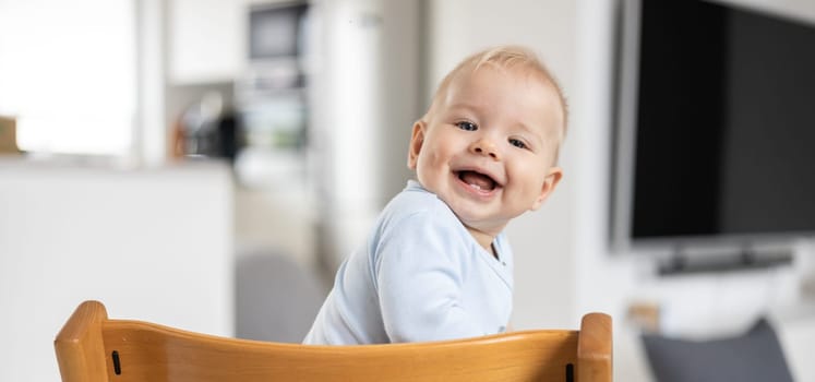 Happy infant sitting in traditional scandinavian designer wooden high chair and laughing out loud in modern bright home. Cute baby smile.