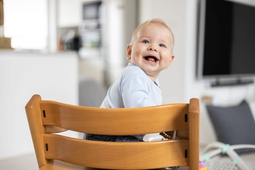 Happy infant sitting in traditional scandinavian designer wooden high chair and laughing out loud in modern bright home. Cute baby smile.