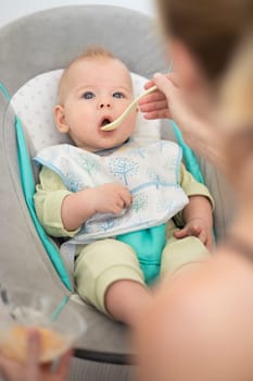 Mother spoon feeding her baby boy infant child in baby chair with fruit puree. Baby solid food introduction concept.