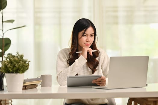 Young asian female worker holding looking at laptop screen, communicating online at workstation