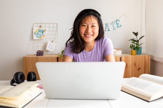 Happy, smiling teen asian female college student studying at home, doing homework using laptop looking at camera.