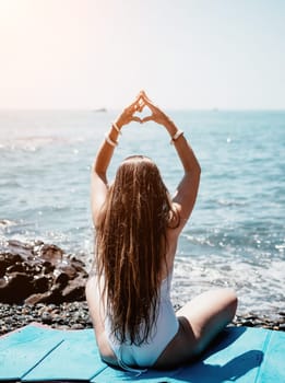 Woman makes heart with hands on beach. Young woman with long hair in white swimsuit and boho style braclets practicing by sea ocean. Concept of longing daydreaming love peace contemplation vacations