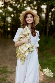 a happy, romantic woman stands in a light dress with a hat on her head and a plaid in her hands in the park