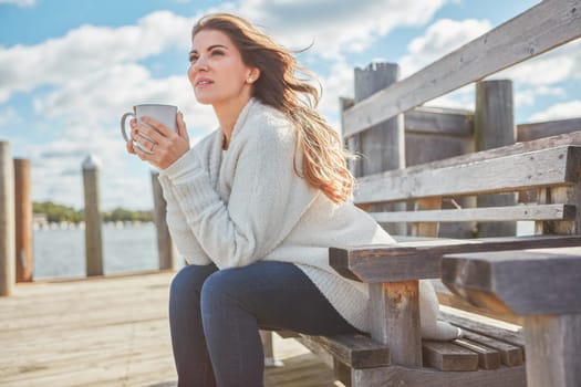 I dont need much - simplicity is the answer for me. a beautiful young woman enjoying a warm beverage while relaxing on a bench at a lake.