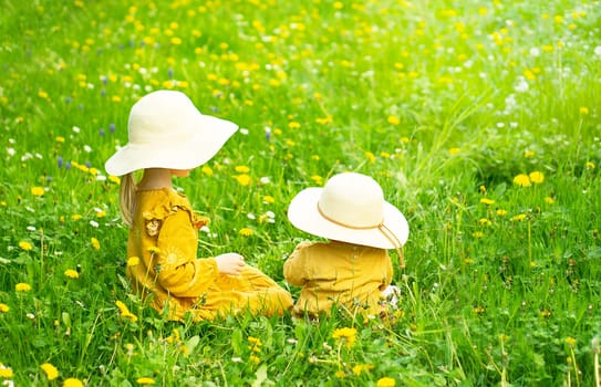 Two sisters in yellow clothes sitting on the green meadow with yellow dandelions.