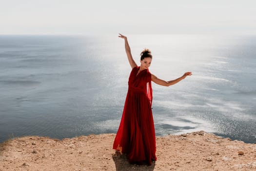 Woman in red dress on sea. Side view a Young beautiful sensual woman in a red long dress posing on a rock high above the sea on sunset. Girl on the nature on blue sky background. Fashion photo.