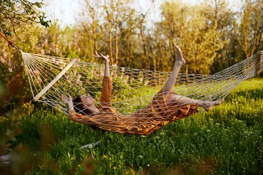 a funny woman is resting in nature lying in a mesh hammock in a long orange dress, lifting up her arms and legs, enjoying the rays of the setting sun