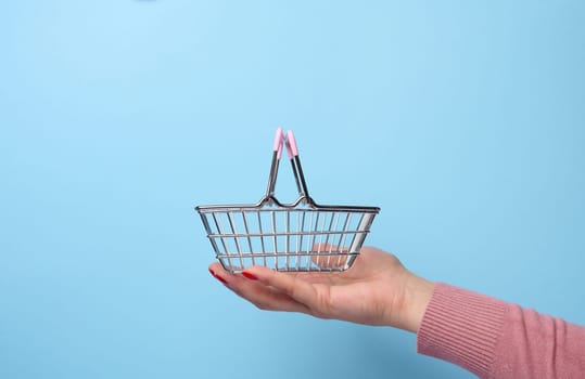 Woman's hand holding a miniature metal shopping basket on a blue background