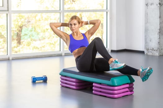 Sporty woman sitting on step platform and doing press aerobic, having work out in fitness club.