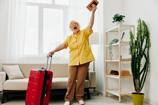 Happy senior woman with passport and travel ticket packed a red suitcase, vacation and health care. Smiling old woman joyfully stands in the house before the trip.