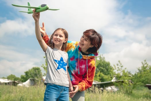 Mother and daughter with toy plane at the field with real airplane on background. Girl and woman smiling looking at each other. Happy family moments at the nature