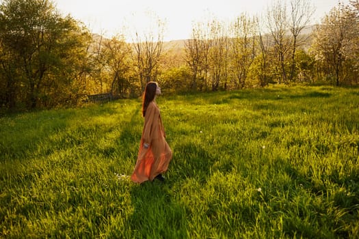 an attractive, slender, red-haired woman stands in a wide green field during sunset in a long orange dress enjoying unity with nature and relaxation