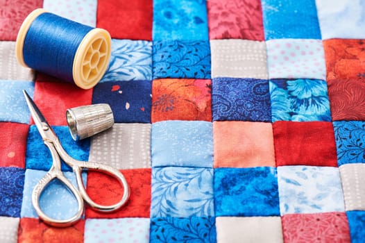 Scissors, thread and thimble lying on blue and red square pieces of fabric sewn together