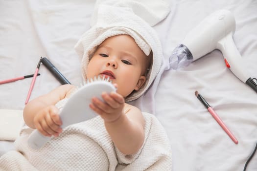 baby with towel on her head lies near the hairdryer and cosmetics