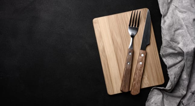 Fork and knife with wooden handle and cutting board on black background, top view