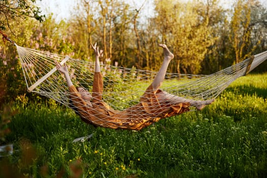 a funny woman is resting in nature lying in a mesh hammock in a long orange dress, lifting up her arms and legs, enjoying the rays of the setting sun