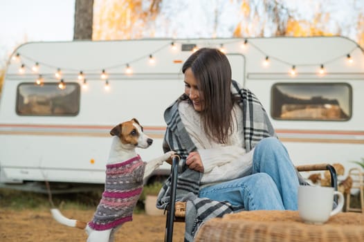 Caucasian woman sitting in a wicker chair wrapped in a blanket with a dog in the yard near the trailer in autumn.