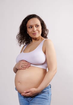 Delightful Hispanic pregnant woman smiling cutely looking aside, caressing her belly, enjoying happy carefree pregnancy