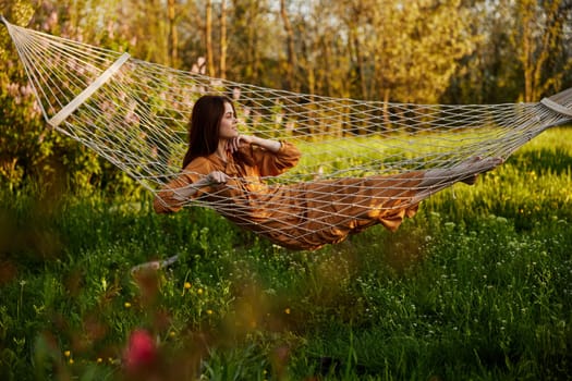 an elegant, slender woman is resting in nature lying in a mesh hammock in a long orange dress enjoying the rays of the setting sun on a warm summer day looking away
