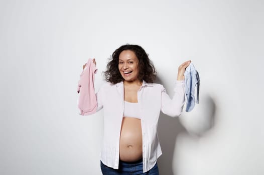 Happy pregnant woman with curly hair, holds pink and blue newborn clothes, expecting twins boy and girl, white backdrop