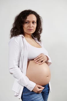 Vertical portrait beautiful pregnant Hispanic woman expecting a baby, posing with hands on naked belly, white background
