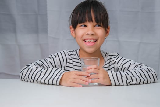 Cute little girl drinking water from glass on white curtain background. Healthy lifestyle and refreshment concept.