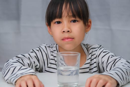 Cute little girl drinking water from glass on white curtain background. Healthy lifestyle and refreshment concept.