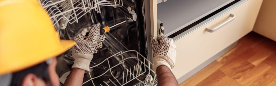 Close up of handyman in special clothing repairing dishwasher in modern kitchen