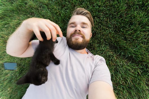 Man taking selfie with smartphone with little kitten lying and playing on grass - friendship love animals and pet owner concept