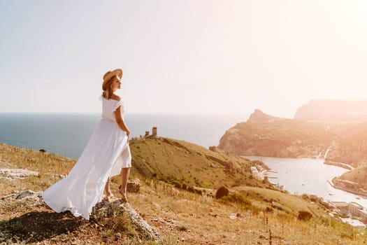 Happy woman in a white dress and hat stands on a rocky cliff above the sea, with the beautiful silhouette of hills in thick fog in the background.