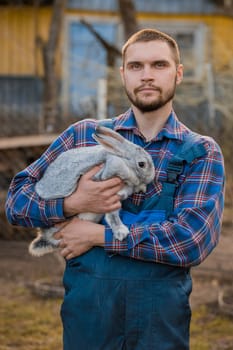 Farmer handsome serious european caucasian rural portrait in countryside with beard, shirt and overalls looking at camera with white rabbit in his arms outdoors