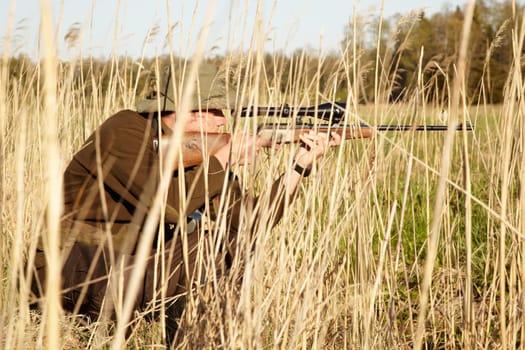 Nature, hunter and man with a rifle while in camouflage shooting in outdoor field. Grass, wildlife and male sniper hunting animals with shotgun weapon hiding in plants to shoot target in countryside.