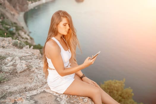 Happy woman in white shorts and T-shirt, with long hair, talking on the phone while enjoying the scenic view of the sea in the background.