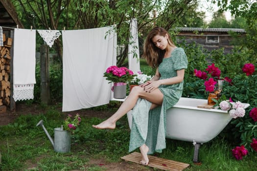 A woman is sitting on a cast-iron bathtub in the courtyard of a country house next to a bush of flowering peonies. The concept of summer, country life, a bathroom on the street in a blooming garden in the country.