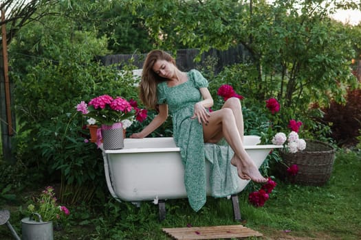 A woman is sitting on a cast-iron bathtub in the courtyard of a country house next to a bush of flowering peonies. The concept of summer, country life, a bathroom on the street in a blooming garden in the country.