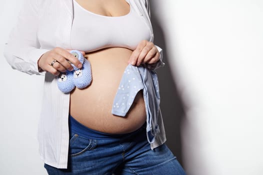 Close-up of gravid woman expecting baby, holding knitted booties and baby bodysuit over her bare belly, white background