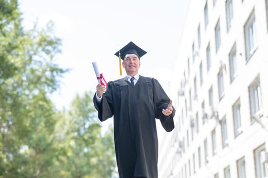 An old man in a graduation gown walks outdoors and holds a diploma.