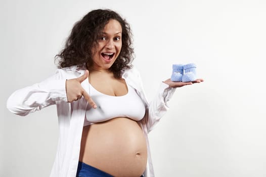 Excited pregnant pretty woman holding baby socks on her hand, pointing at her belly, rejoicing at her pregnancy 30 week