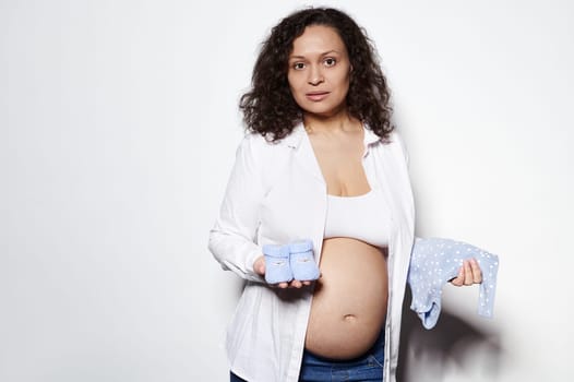 Portrait of pregnant woman on second trimester of pregnancy, posing with blue newborn bodysuit and baby shoes on white