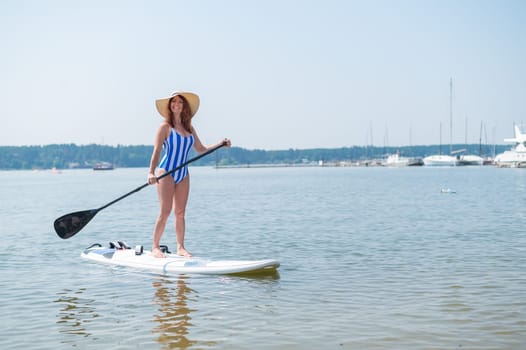 A red-haired woman in a striped swimsuit and a straw hat floats on a paddle board in the yacht club